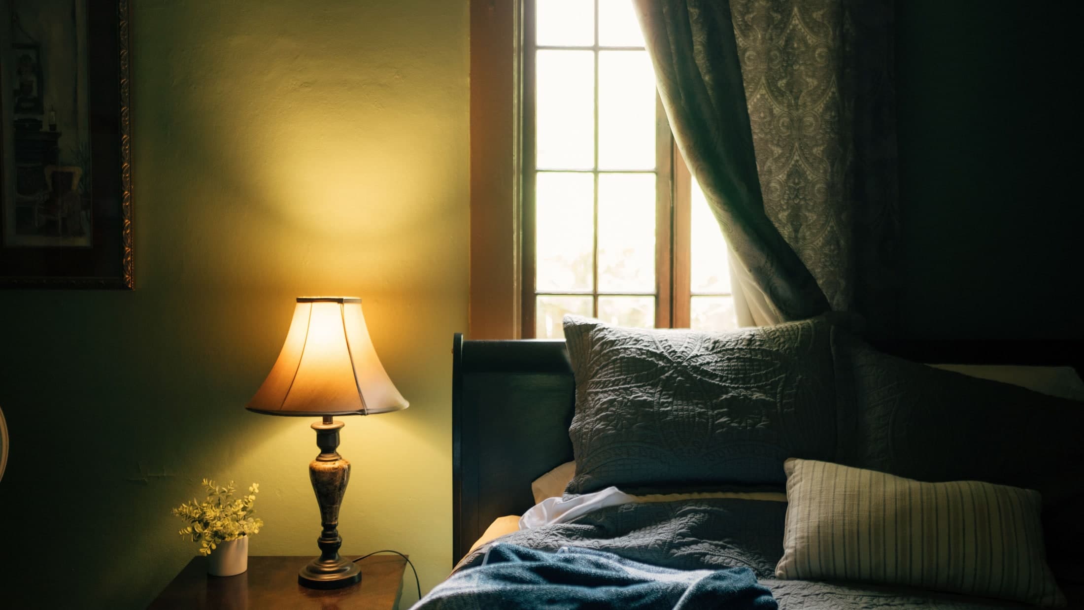 A lit tapered shade stands beside dark patterned bedding, with daylight at the window and a patterned curtain.