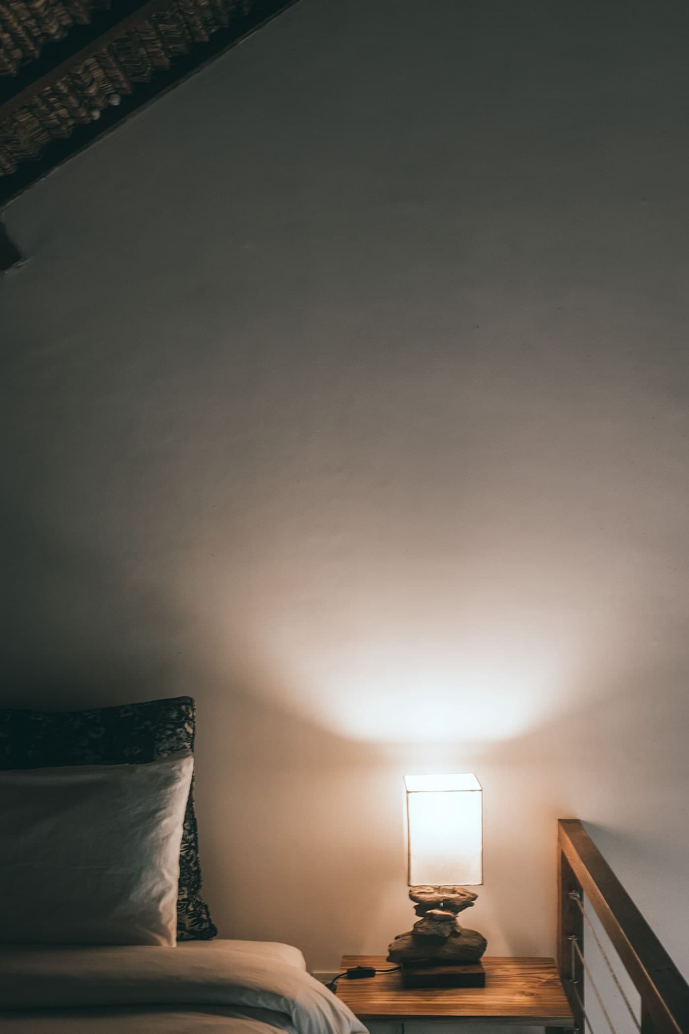 A rectangular glowing shade on an irregular dark base lights the wall above a timber bedside surface, with pillows beside it.
