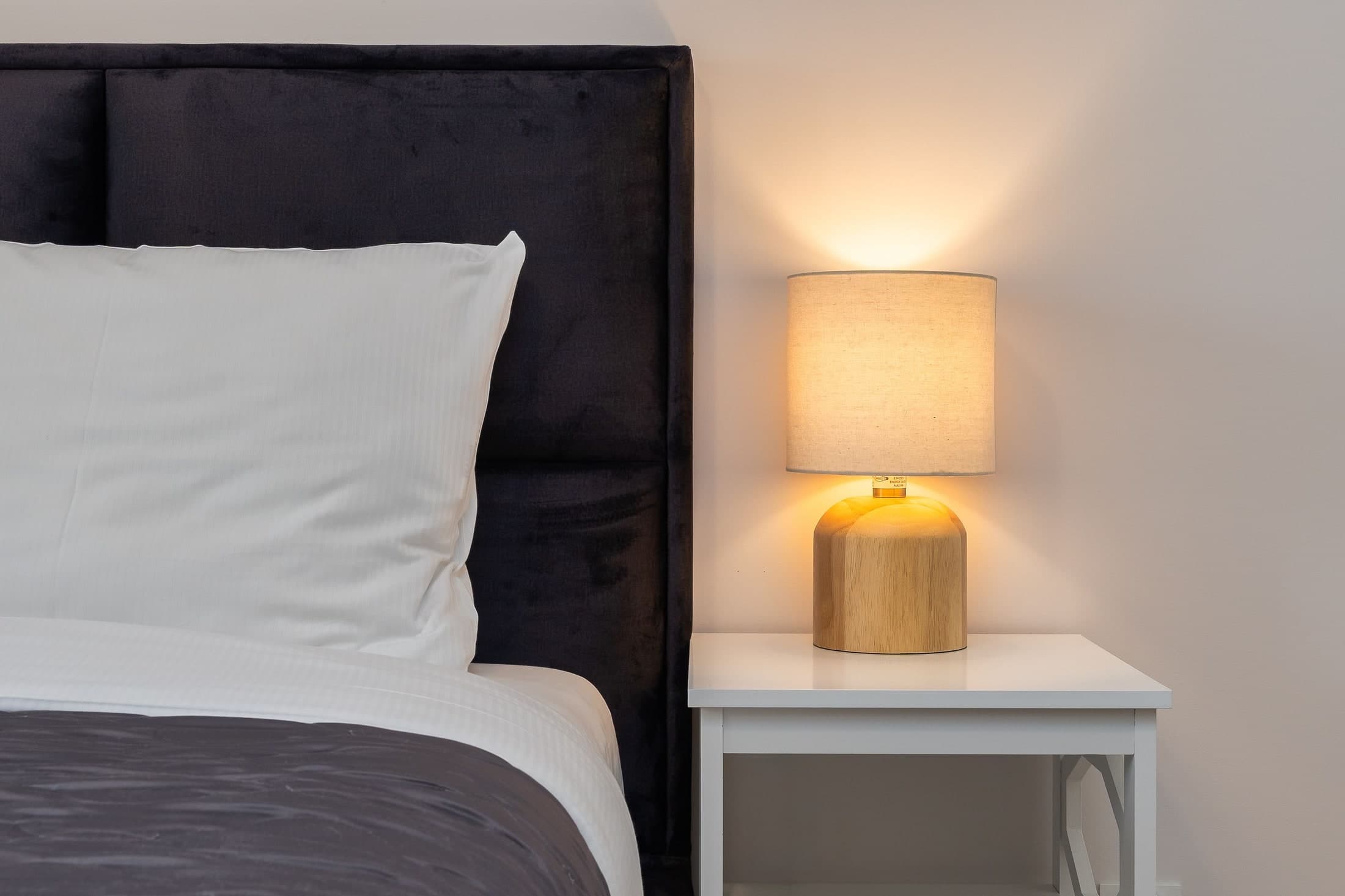 A glowing round shade with a rounded timber-looking base stands on a pale table beside white pillows and a dark headboard.