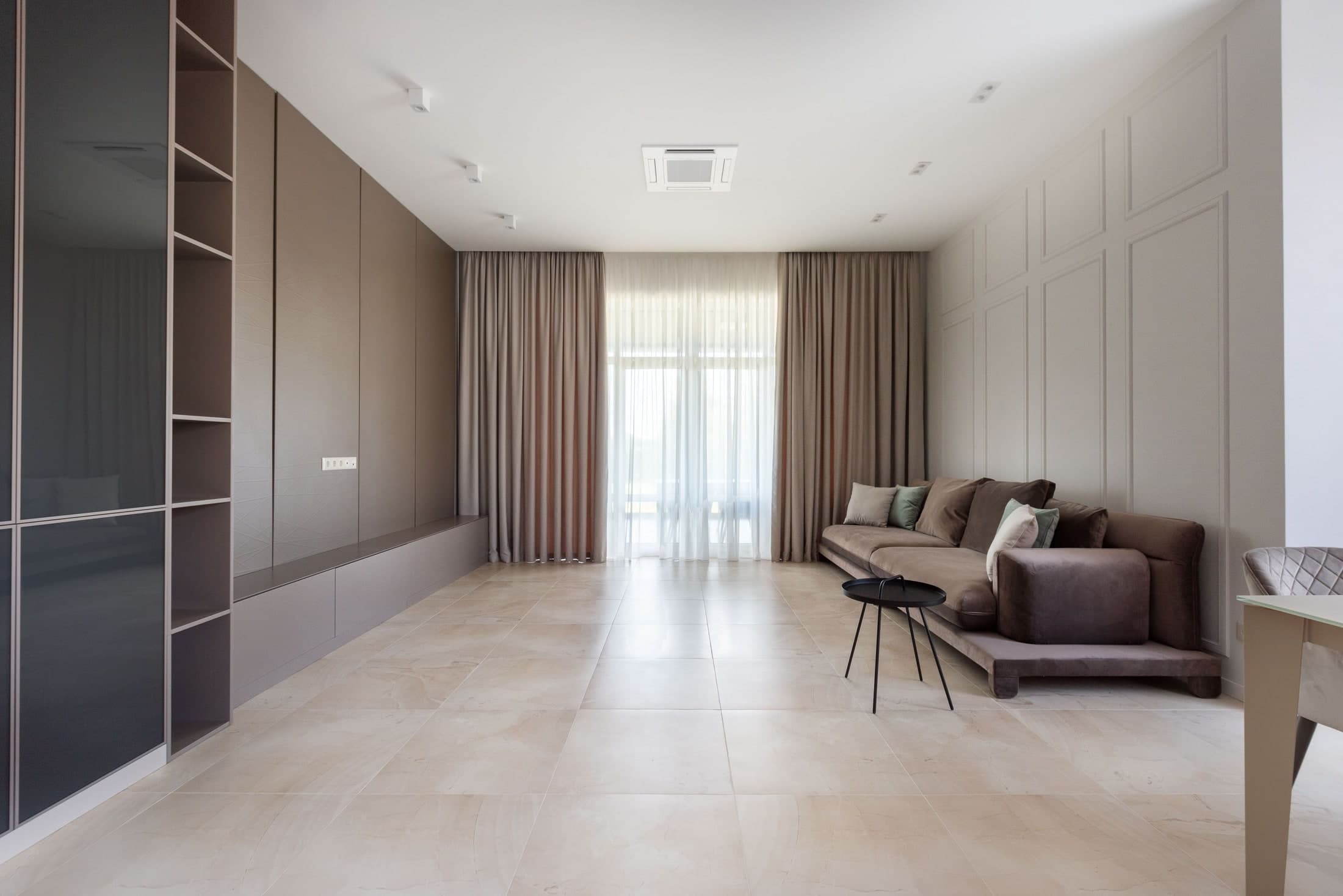 Pale full-length curtains flank a sheer layer across glazing, with a sofa to the right and tall cupboards to the left.
