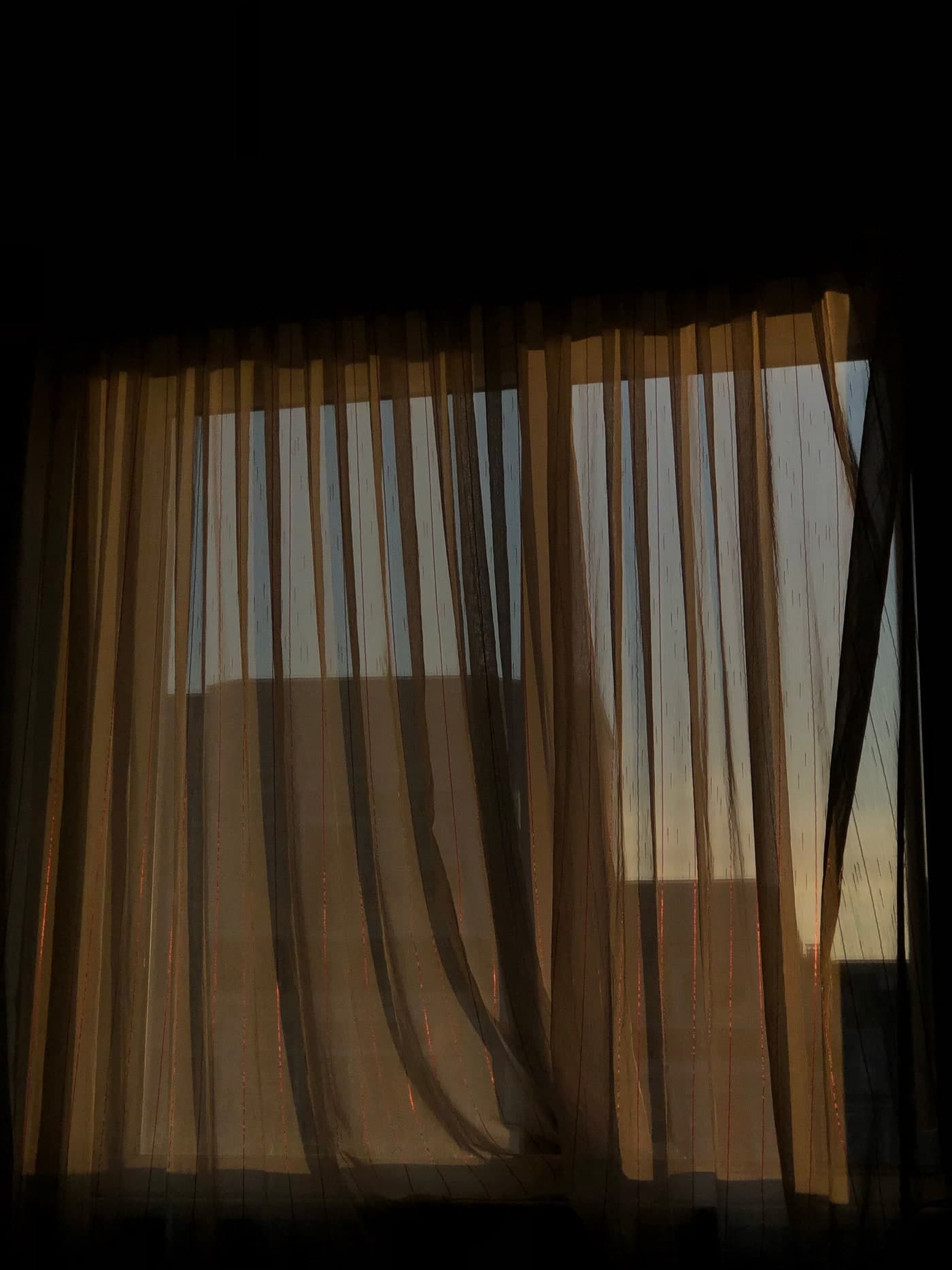 Light passes through fine curtains with narrow vertical lines, leaving the window frame and a building visible beyond.