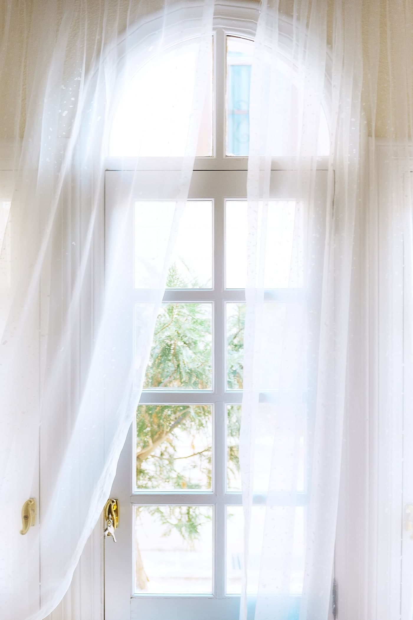 Fine pale curtains hang in loose straight folds across a white arched glazed door with brass-coloured handles.