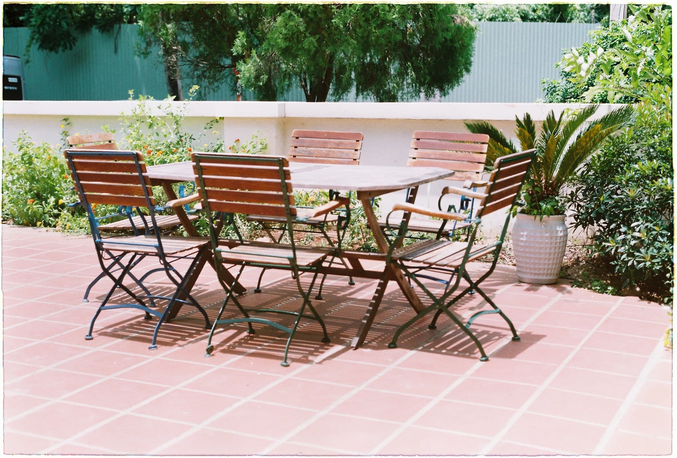 Timber chairs surround an oval table on red tiles beside a pale low wall and green planting.