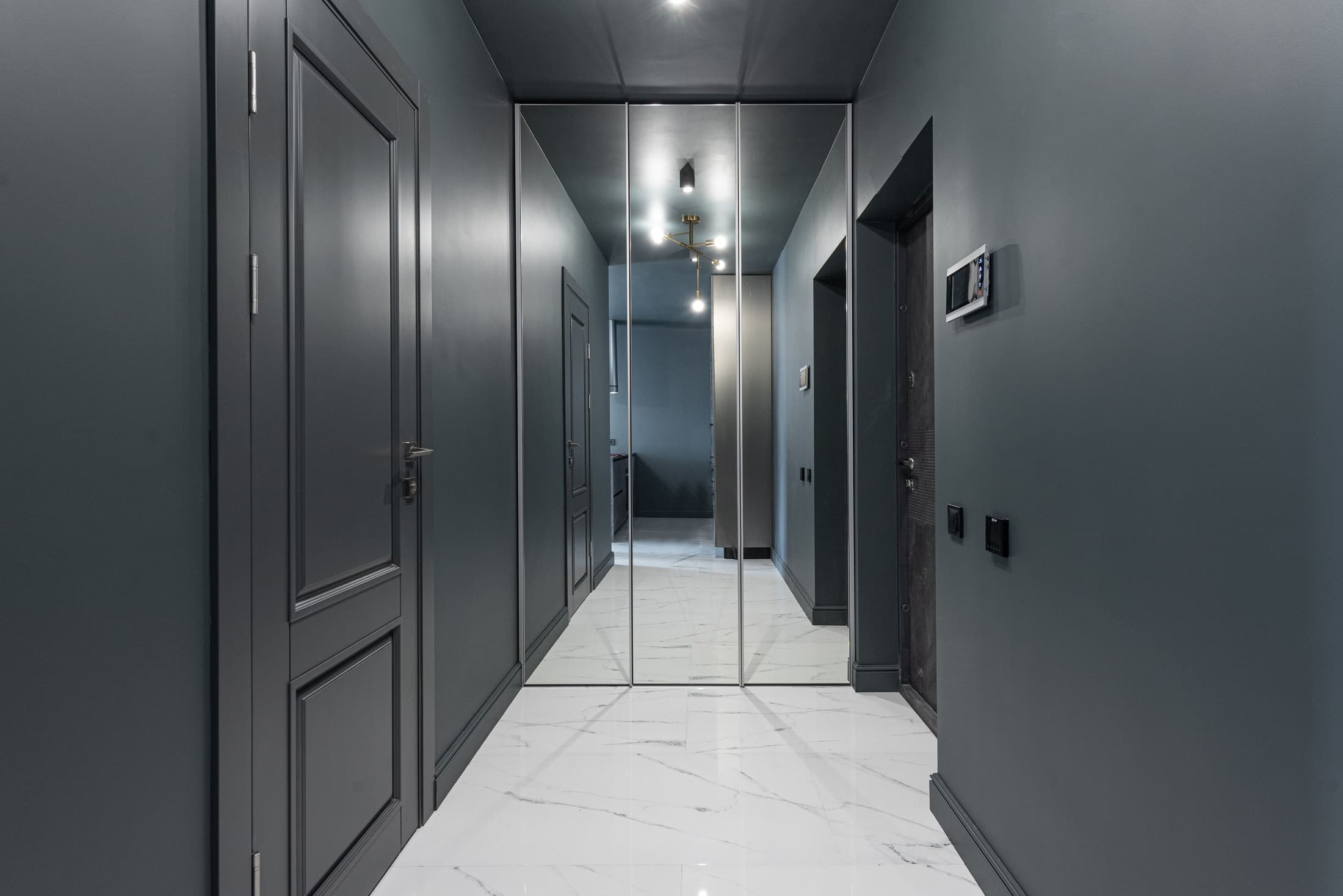 Three tall mirror panels at the end of a dark hallway reflect doors, ceiling lights and a pale veined floor.
