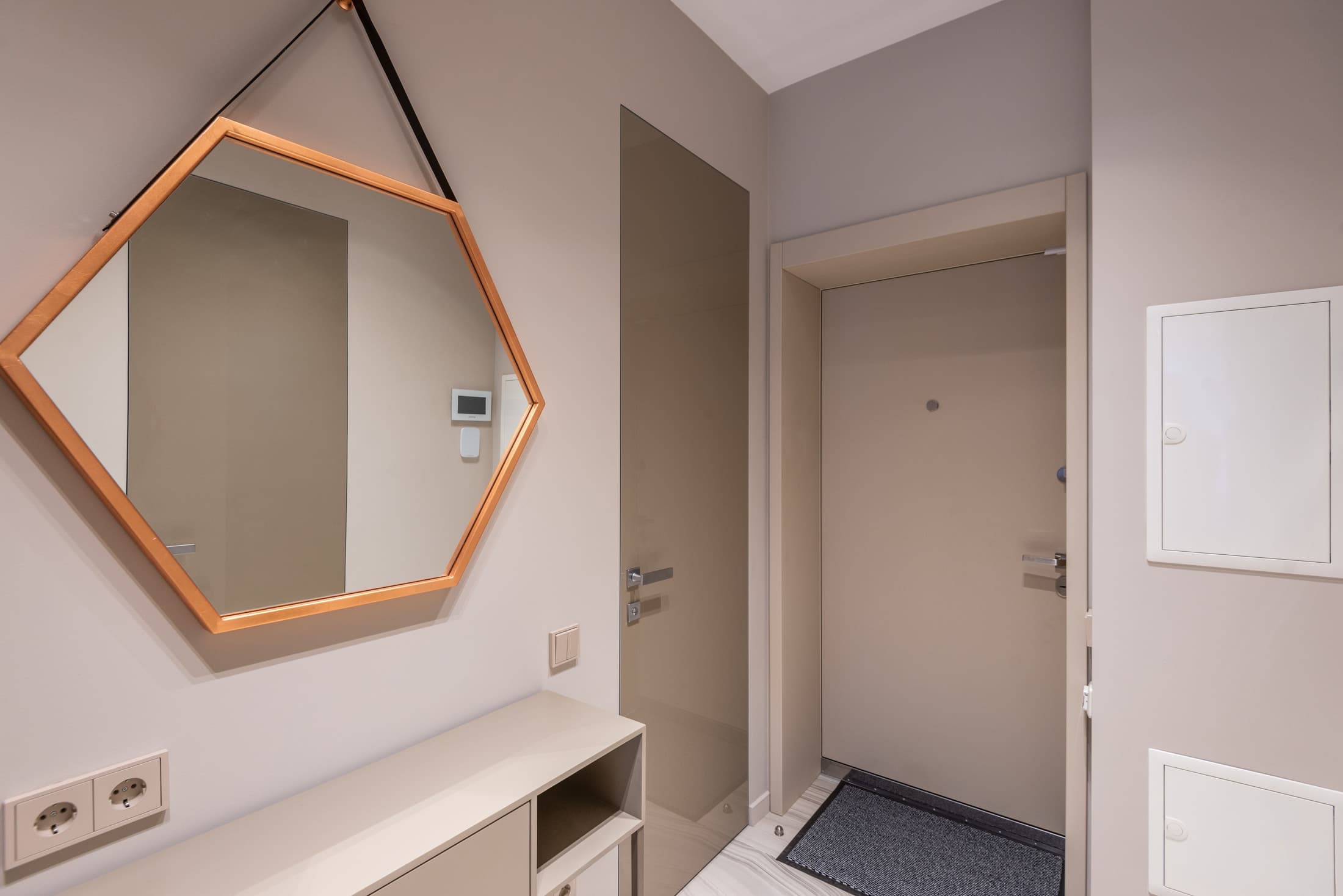 A hexagonal mirror suspended from a dark strap above a pale console reflects a door and wall controls.