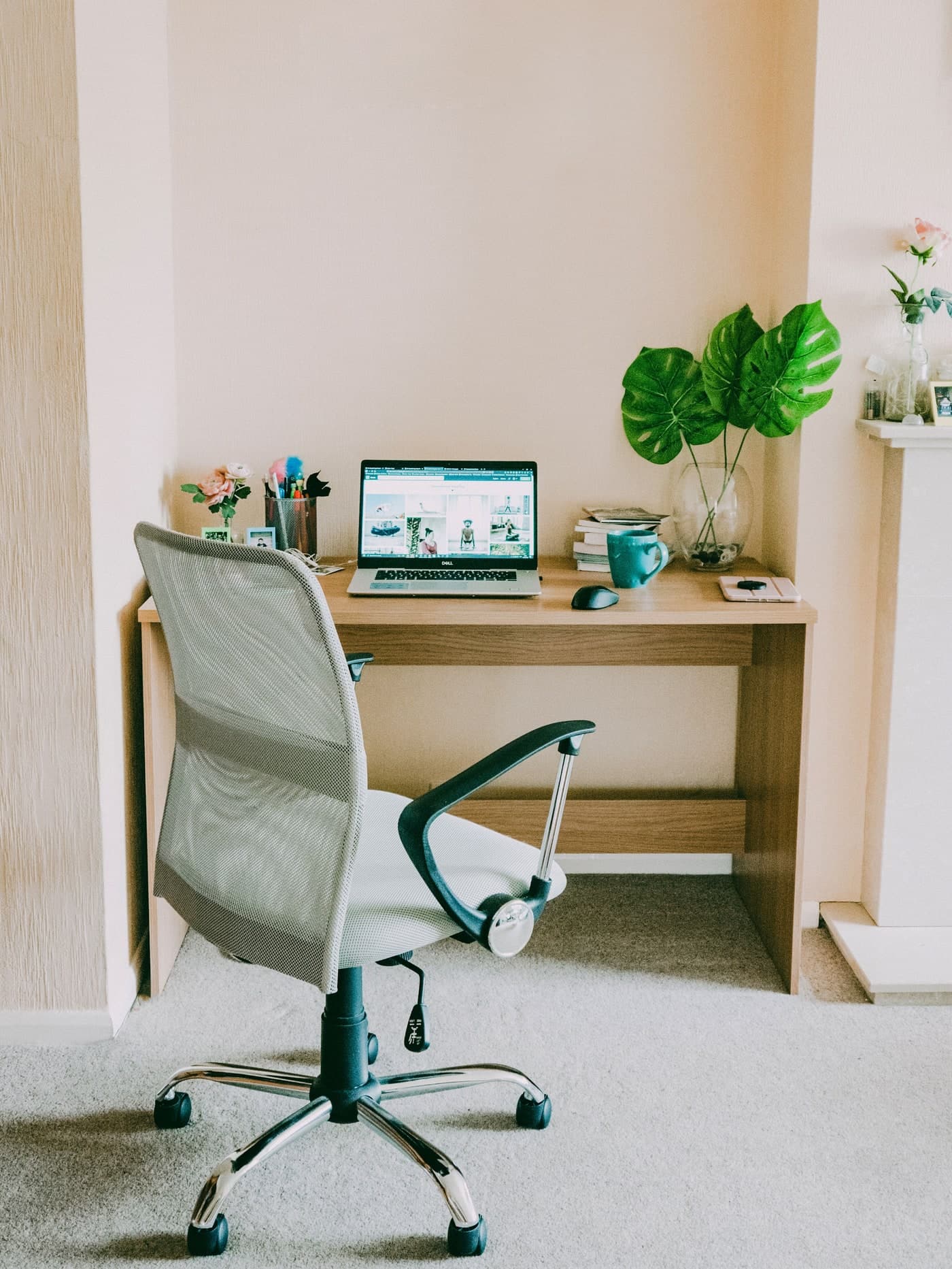 A grey mesh office chair stands at a timber desk with a laptop, mouse, stationery, cup and leafy stems.