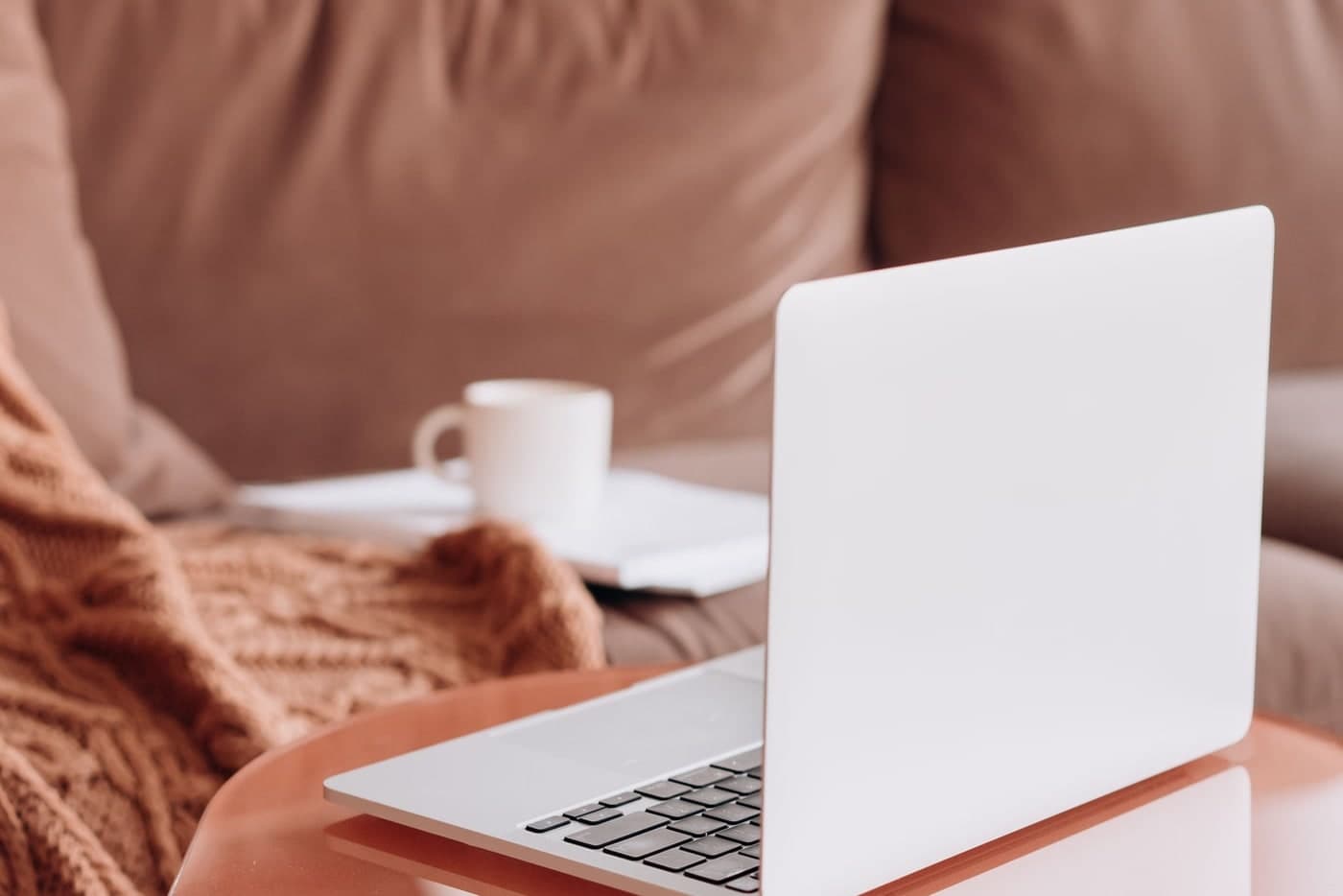 An open laptop rests on an orange tray beside a brown sofa, with a cup and notebook behind it.