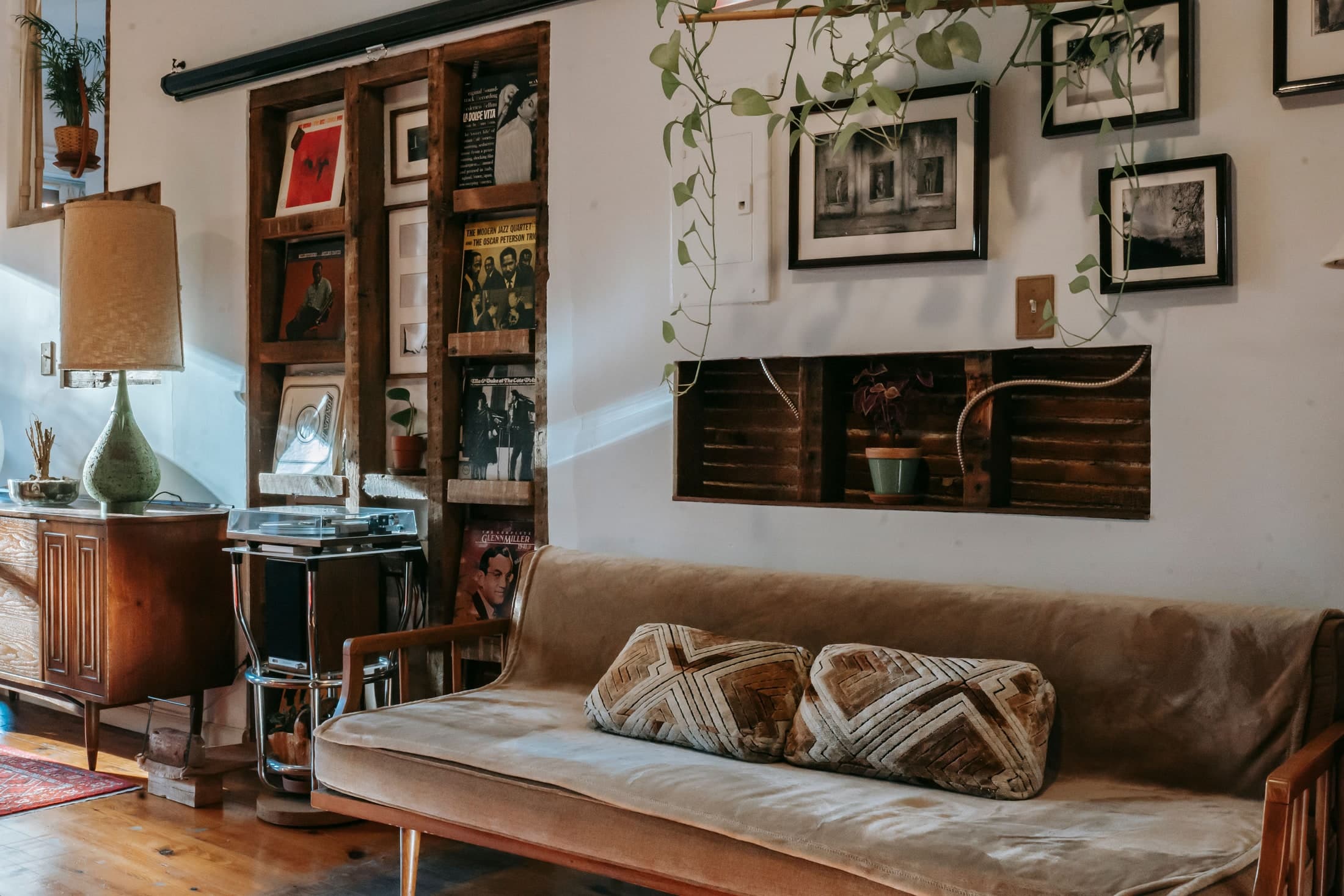 A shiny metal turntable stand sits between a panelled wooden cabinet and a pale-covered timber sofa with patterned cushions.