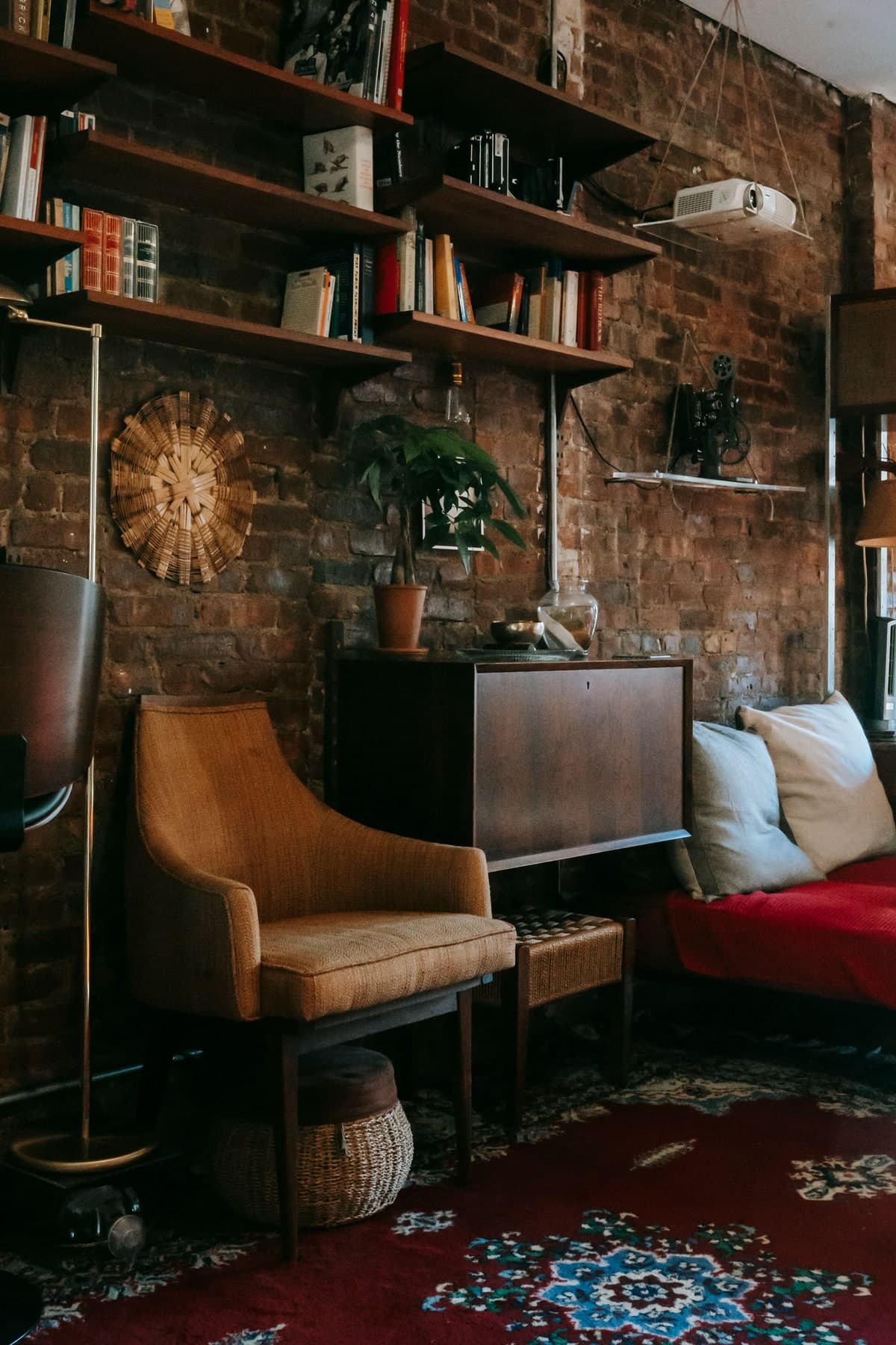 An upholstered timber armchair sits beneath bookshelves on an exposed brick wall, beside a dark wooden cabinet.