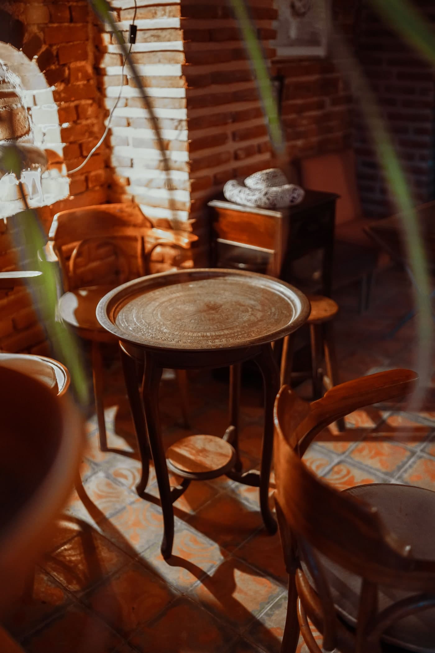 Timber chairs and a stool sit around a round tray-topped table, with reddish brickwork and patterned floor tiles.