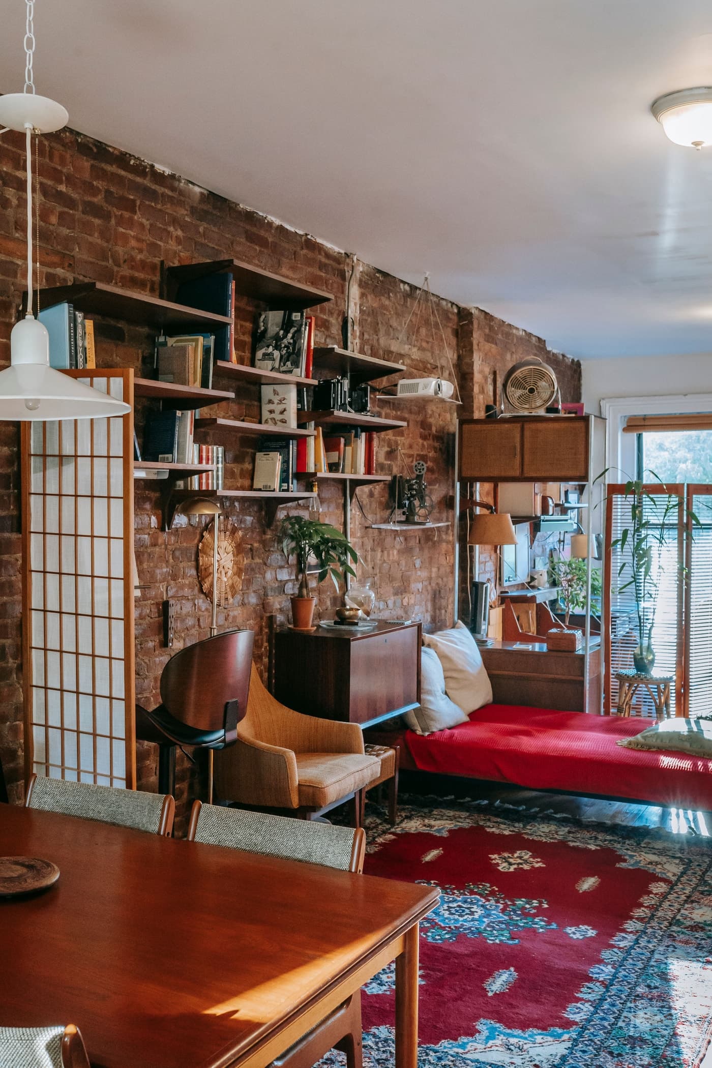 A red patterned rug extends towards a timber dining table, beside a timber-framed armchair and red-covered seat in a brick-walled room.
