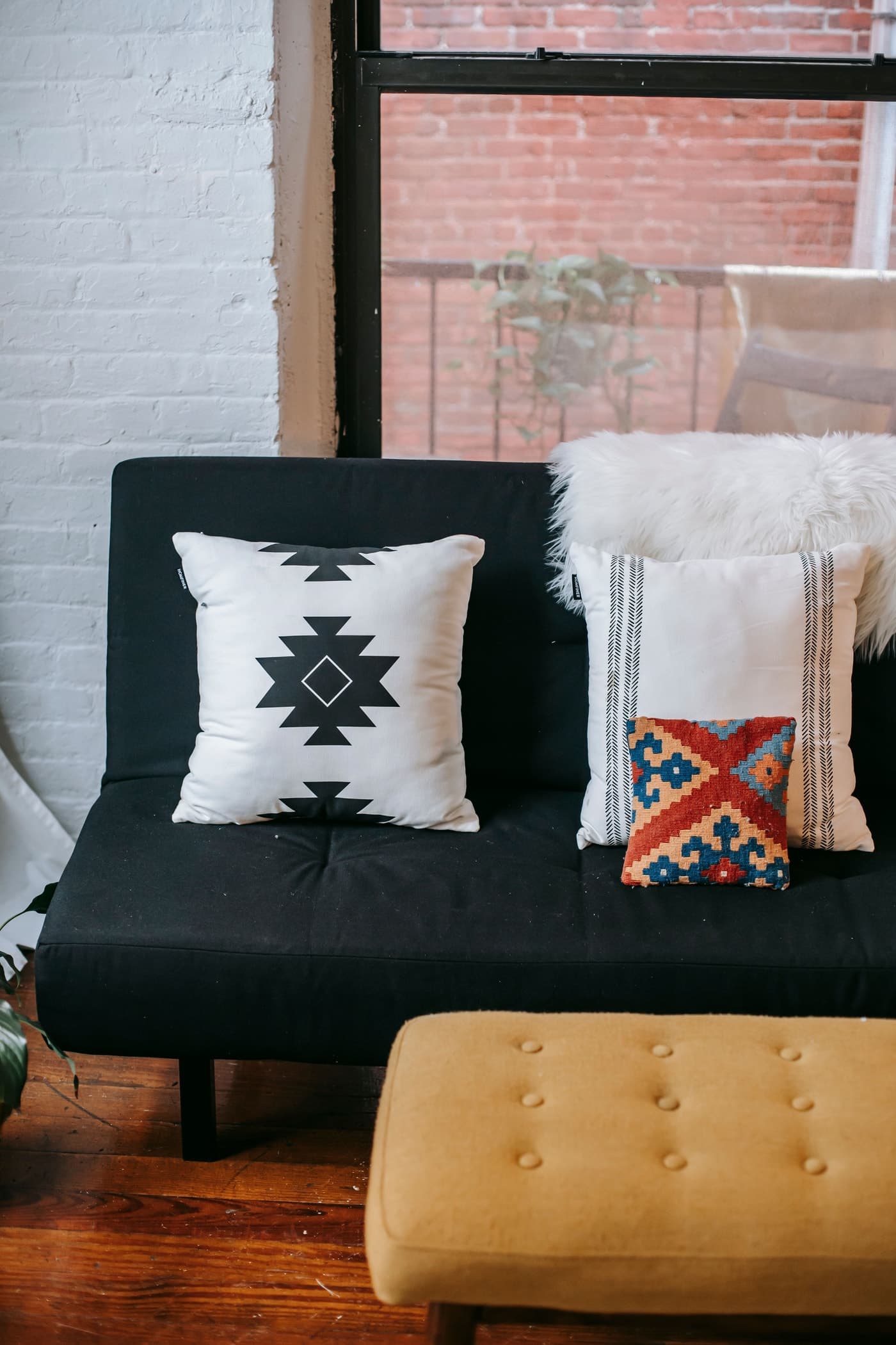 A dark upholstered seat and mustard footstool stand on timber boards beneath a window, with white and patterned cushions.