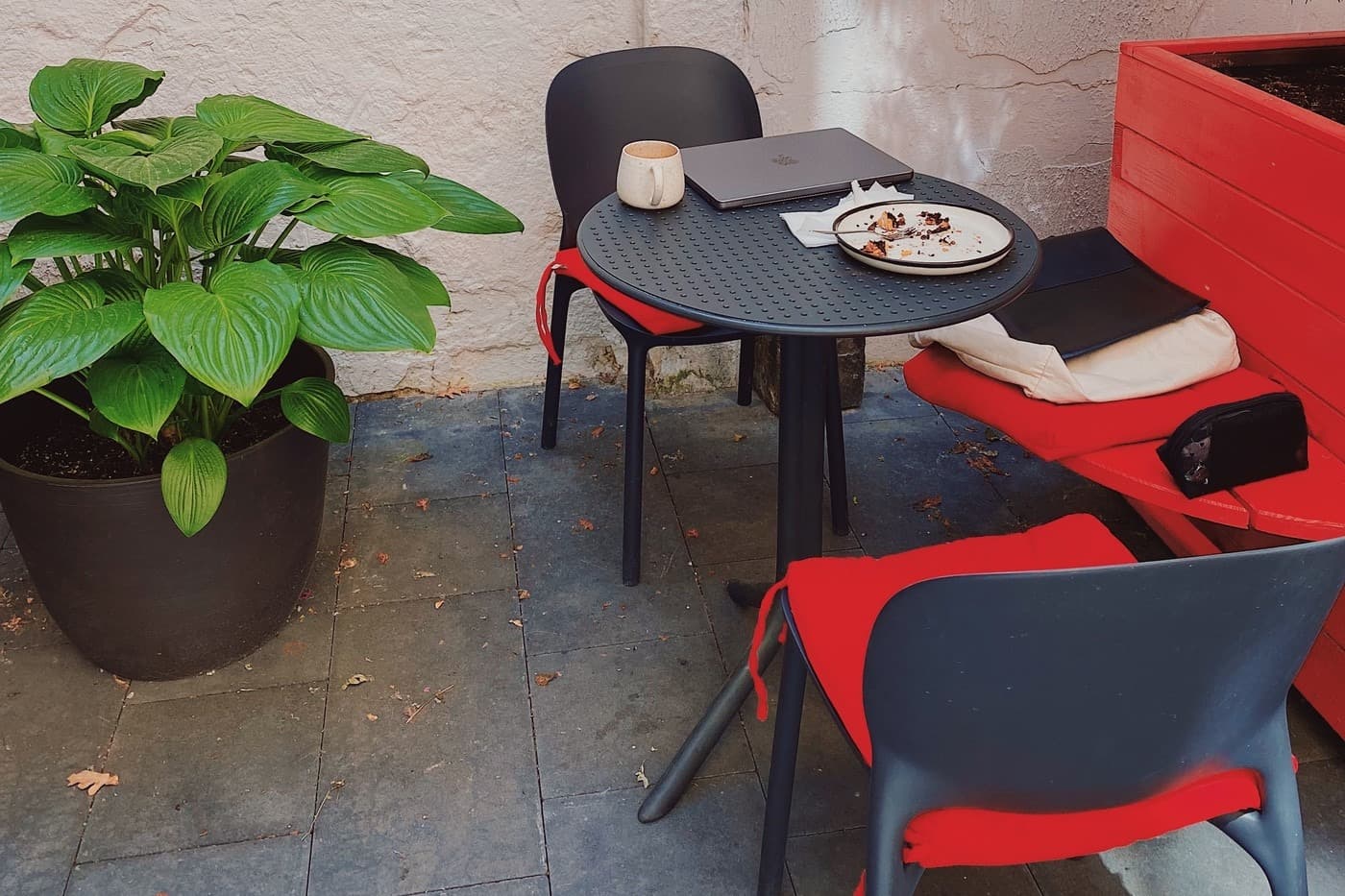 A round black table with a laptop, mug and plate is surrounded by dark chairs with red cushions, between a large plant pot and a red planter.