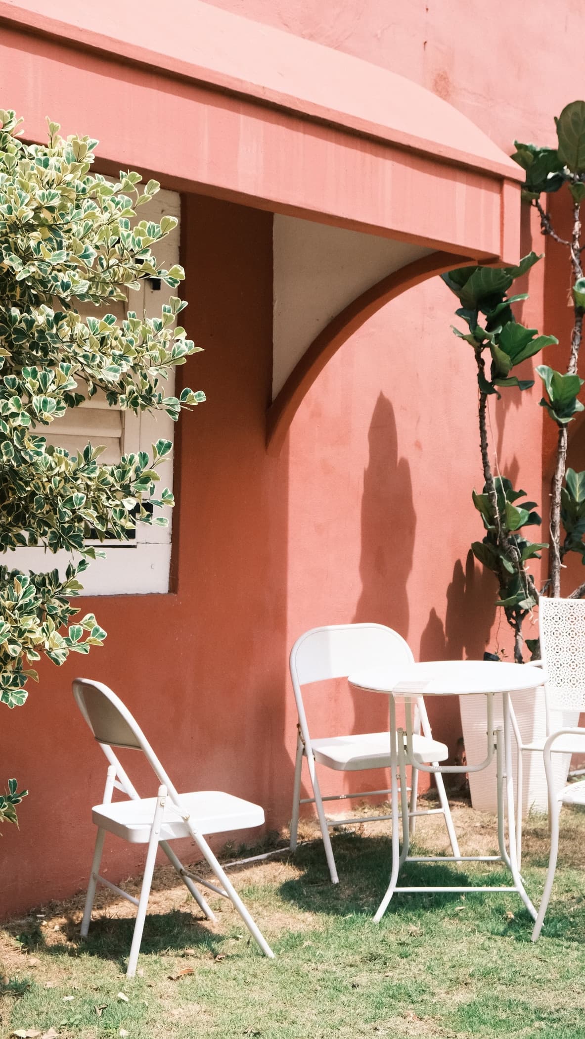 White chairs and a round white table stand on grass against a pink wall, with foliage on either side.