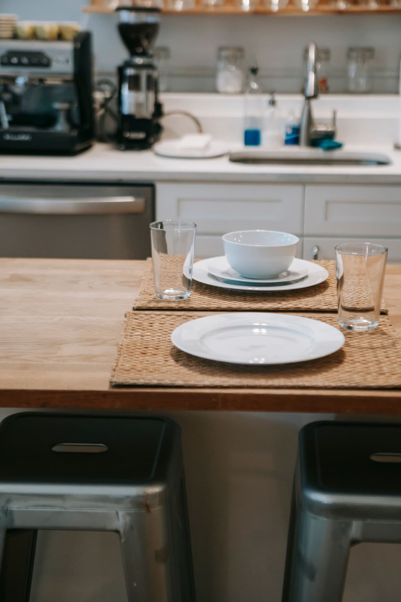 Two dark stools below a timber surface set with woven placemats, plates, bowls and glasses; a sink and coffee appliances stand behind.