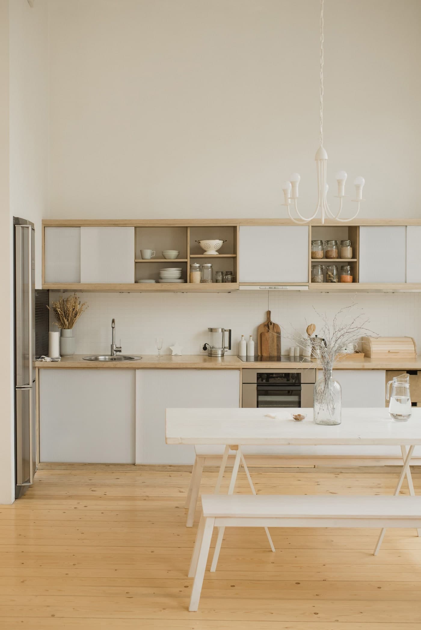 A long pale trestle table and bench stand before kitchen cupboards, with an open fridge door at the left.