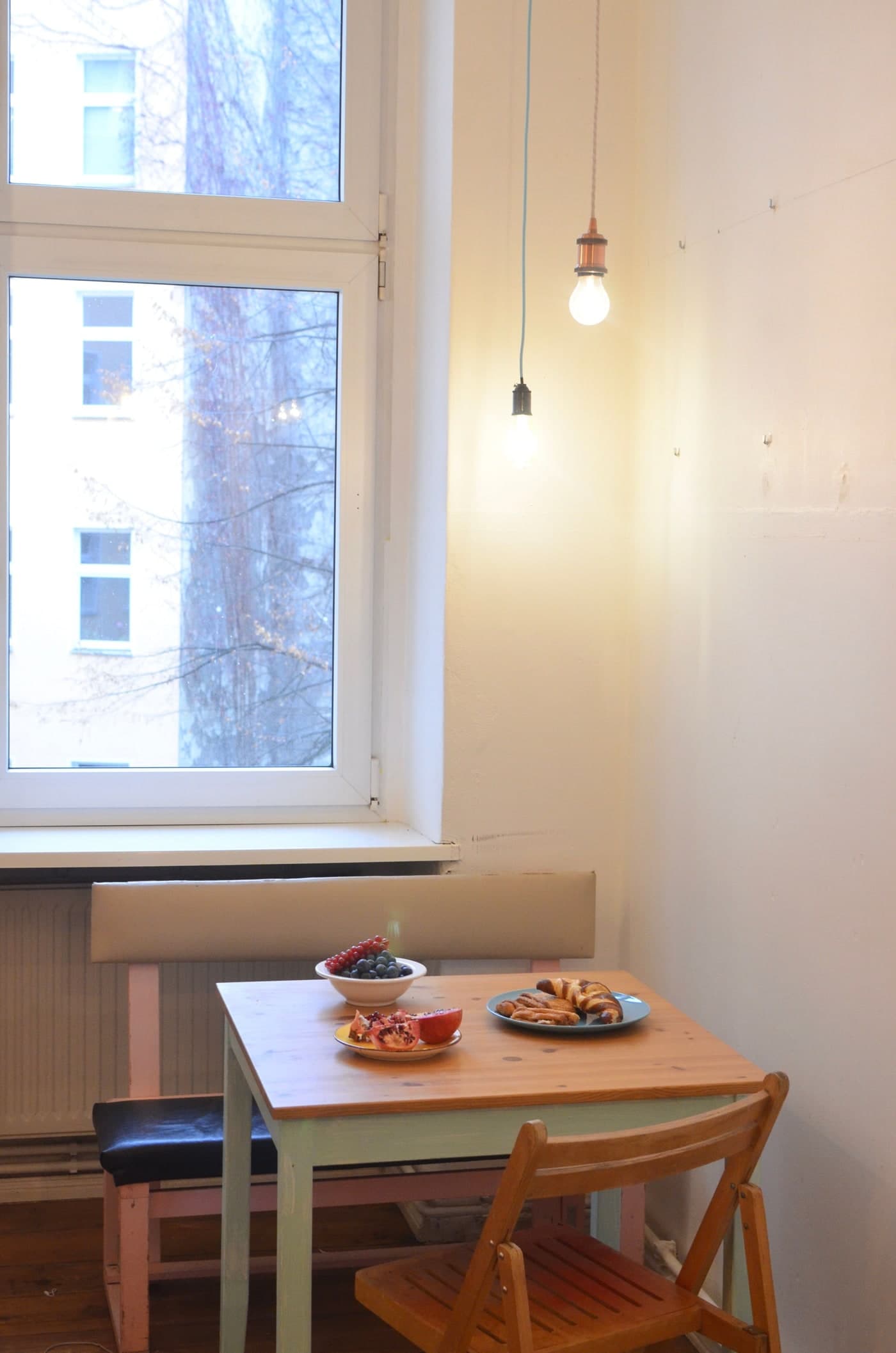 A pale-green timber-topped table with fruit and pastries, a loose backed bench behind and a wooden chair in front, beneath two lit pendant bulbs.