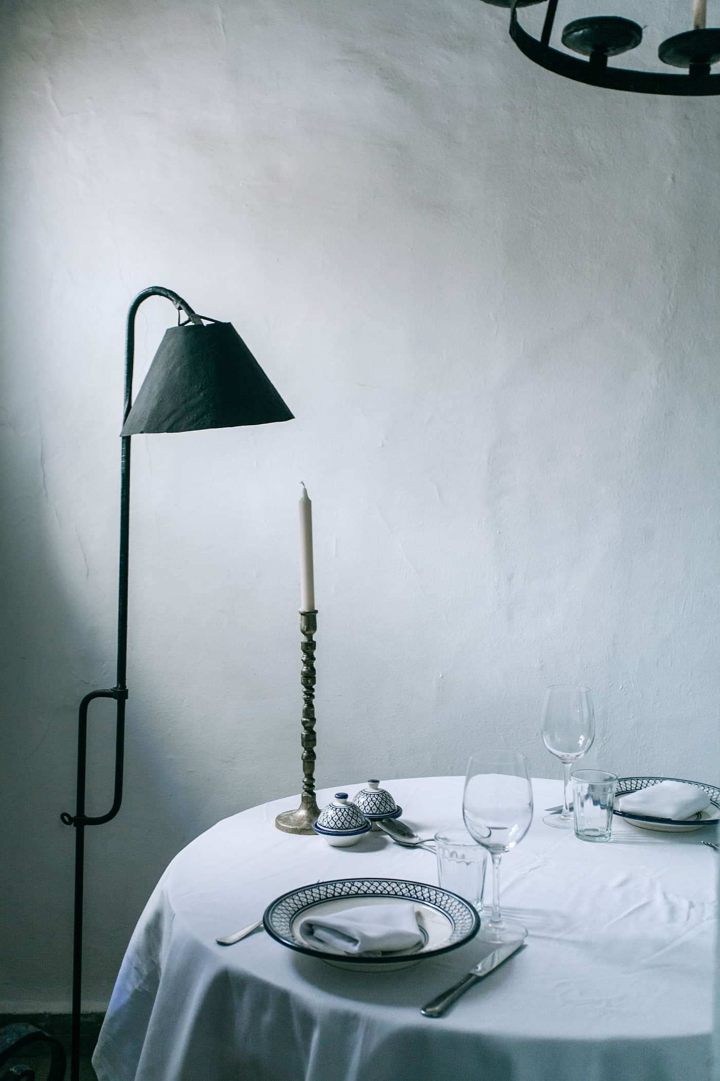 A table with a pale cloth, glasses, patterned crockery and an unlit candle stands beside a dark lamp and textured pale wall.